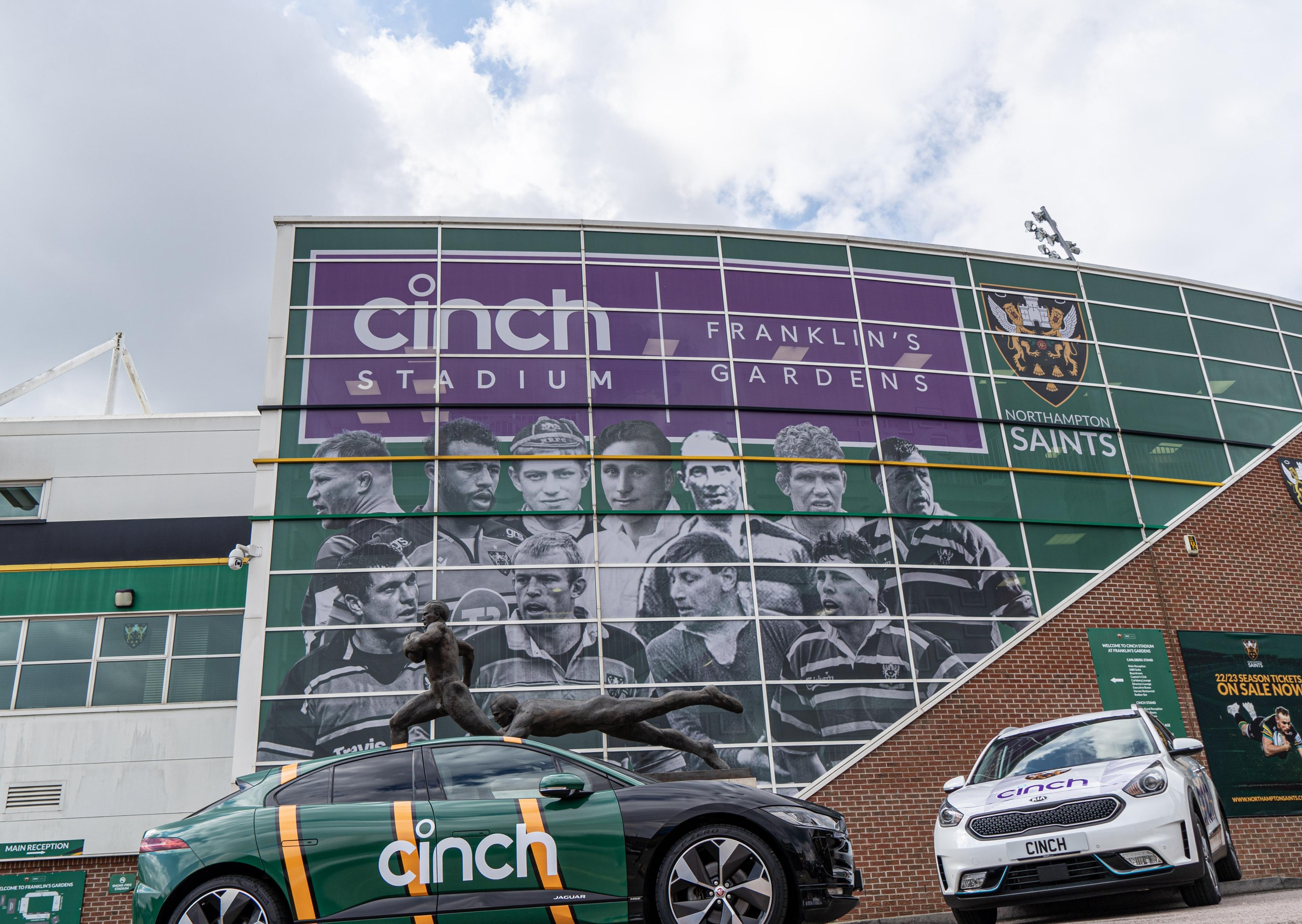 Two cars with "cinch" branding parked in front of a stadium featuring a large mural of rugby players and the "Franklin's Gardens" sign.