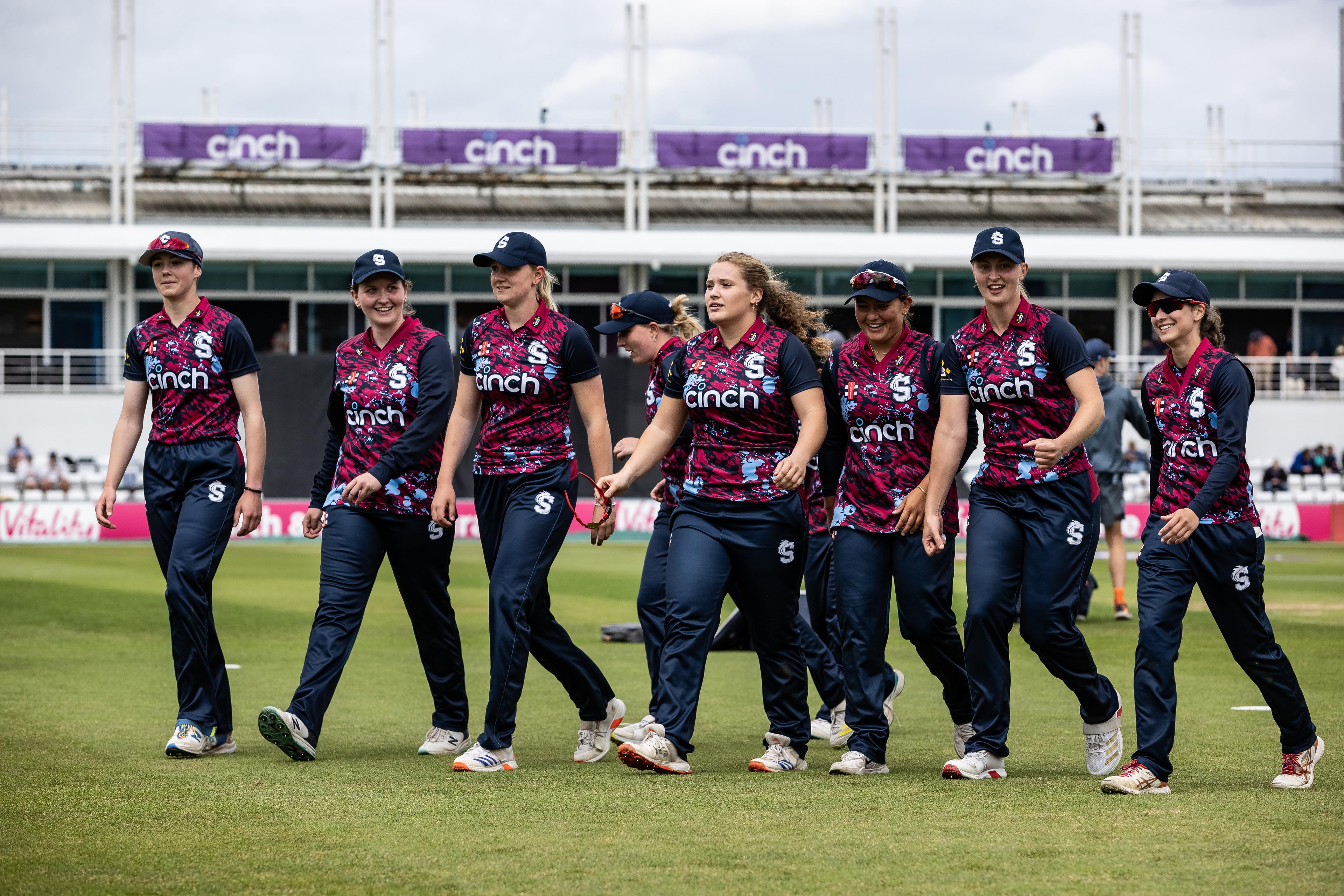 A women's cricket team in matching uniforms walks together on a grassy field, with a stadium and cloudy sky in the background.