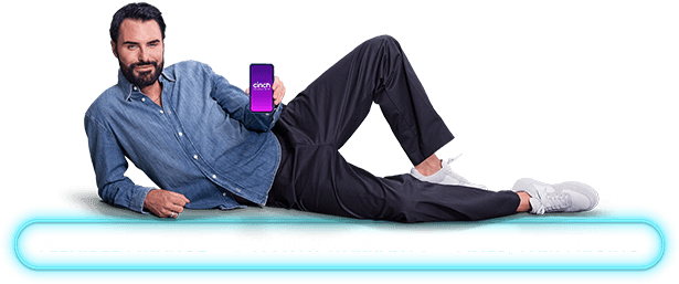Man in a denim shirt and dark pants lies on a turquoise surface, holding a smartphone displaying a purple screen with the word "enjoy."