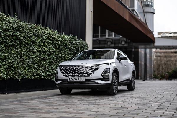 A modern white OMODA 5 SUV with a distinctive grille is parked on a cobblestone street, next to a building with green ivy-covered walls.